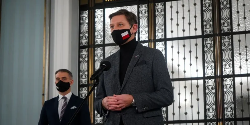 Dwóch mężczyzn w garniturach stoi przy mikrofonie podczas konferencji prasowej w eleganckim wnętrzu z ozdobnymi metalowymi drzwiami w tle; jeden wygłasza przemówienie, drugi stoi za nim.