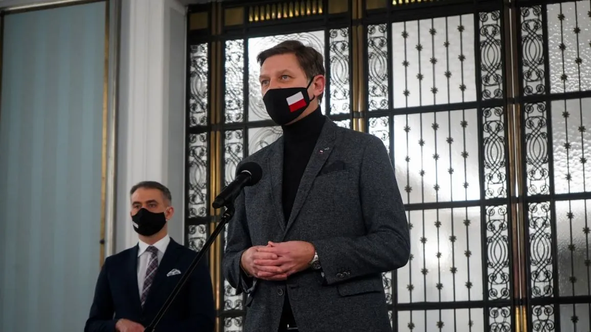 Dwóch mężczyzn w garniturach stoi przy mikrofonie podczas konferencji prasowej w eleganckim wnętrzu z ozdobnymi metalowymi drzwiami w tle; jeden wygłasza przemówienie, drugi stoi za nim.