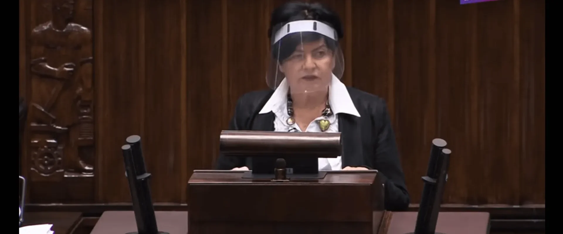 Kobieta w czarnym garniturze z białą koszulą stoi za mównicą podczas posiedzenia, nosi przyłbicę ochronną, a w tle widać drewniane panele i logotyp Lewica w rogu.