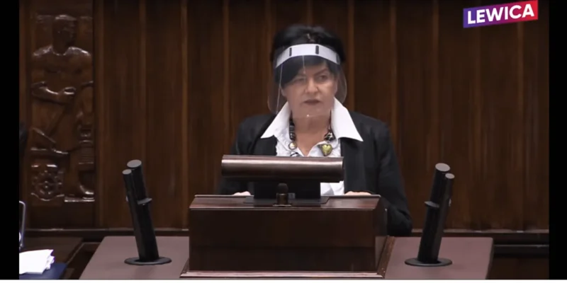 Kobieta w czarnym garniturze z białą koszulą stoi za mównicą podczas posiedzenia, nosi przyłbicę ochronną, a w tle widać drewniane panele i logotyp Lewica w rogu.