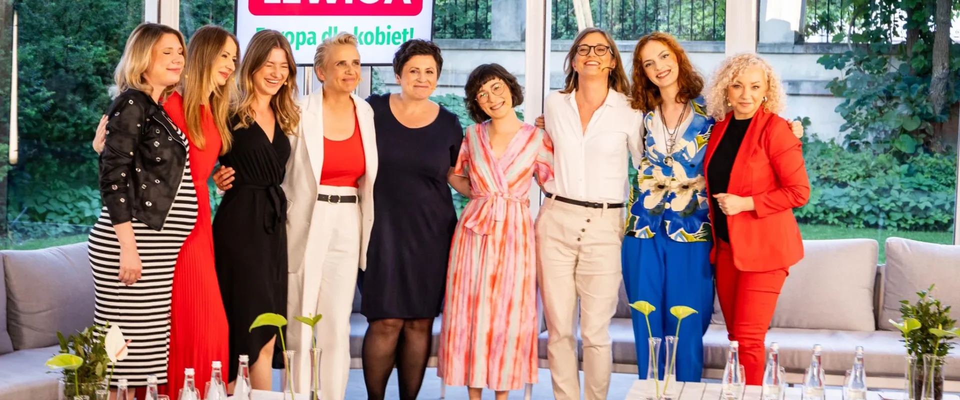 Grupa dziewięciu kobiet stoi razem na tle baneru Lewicy z napisem „Europa dla kobiet”, podczas konwencji programowej, uśmiechnięta i ubrana w kolorowe stroje.