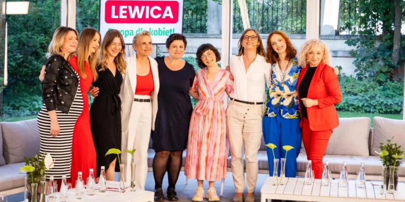 Grupa dziewięciu kobiet stoi razem na tle baneru Lewicy z napisem „Europa dla kobiet”, podczas konwencji programowej, uśmiechnięta i ubrana w kolorowe stroje.