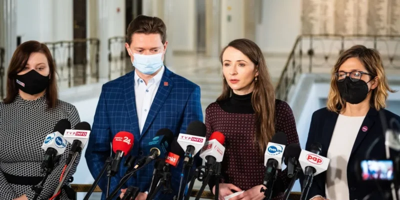 Czworo polityków w maskach ochronnych podczas konferencji prasowej w budynku z jasnym wnętrzem i mikrofonami przed nimi.