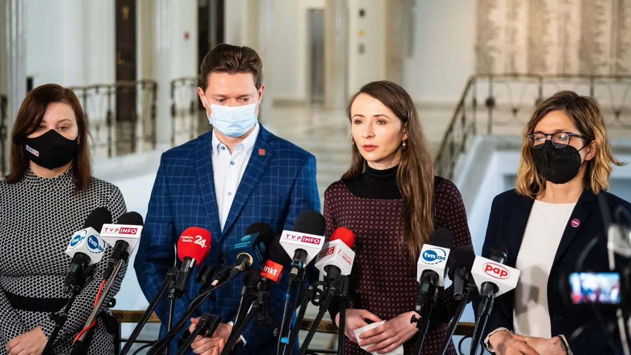 Czworo polityków w maskach ochronnych podczas konferencji prasowej w budynku z jasnym wnętrzem i mikrofonami przed nimi.