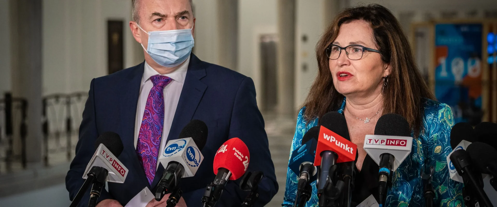 Dwóch polityków stoi przy mikrofonach podczas konferencji prasowej o wzroście cen energii; mężczyzna w granatowym garniturze i maseczce oraz kobieta w okularach i turkusowej bluzce rozmawiają z mediami.