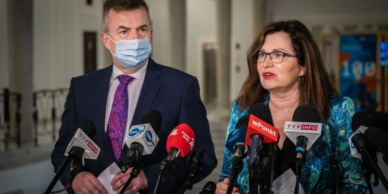 Dwóch polityków stoi przy mikrofonach podczas konferencji prasowej o wzroście cen energii; mężczyzna w granatowym garniturze i maseczce oraz kobieta w okularach i turkusowej bluzce rozmawiają z mediami.