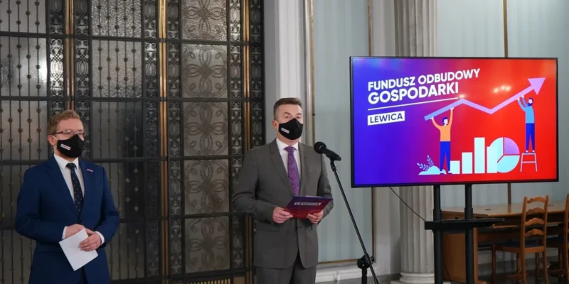 Dwóch mężczyzn w garniturach i maseczkach stoi przy mikrofonie podczas prezentacji o Funduszu Odbudowy Gospodarki; na ekranie w tle widać napis „Fundusz Odbudowy Gospodarki” i rosnący wykres.