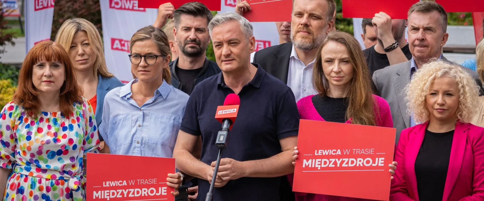Grupa polityków Lewicy podczas konferencji prasowej w Międzyzdrojach, trzymająca czerwone plakaty z napisem LEWICA i stojąca wokół mikrofonu.
