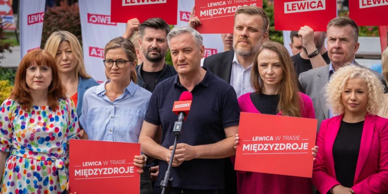 Grupa polityków Lewicy podczas konferencji prasowej w Międzyzdrojach, trzymająca czerwone plakaty z napisem LEWICA i stojąca wokół mikrofonu.