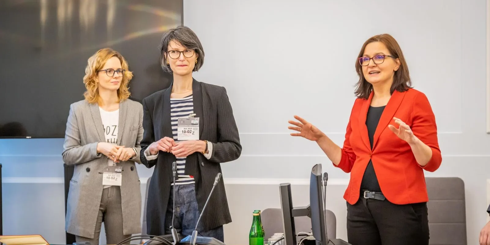 Trzy kobiety w profesjonalnym otoczeniu dyskutują, jedna z nich prowadzi prezentację, a w tle znajdują się nowoczesne urządzenia biurowe.