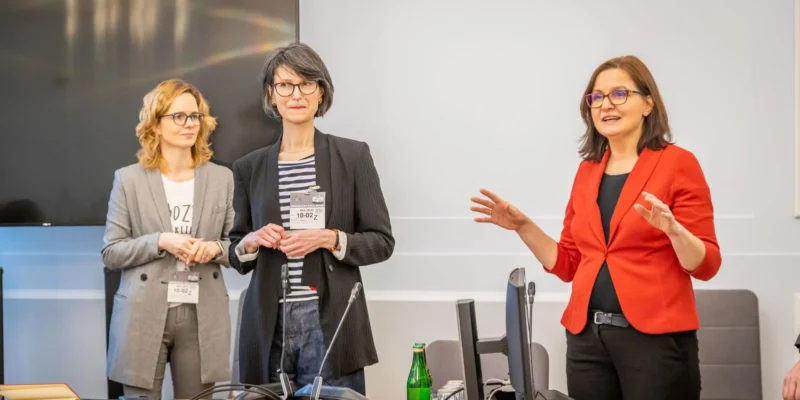 Trzy kobiety w profesjonalnym otoczeniu dyskutują, jedna z nich prowadzi prezentację, a w tle znajdują się nowoczesne urządzenia biurowe.