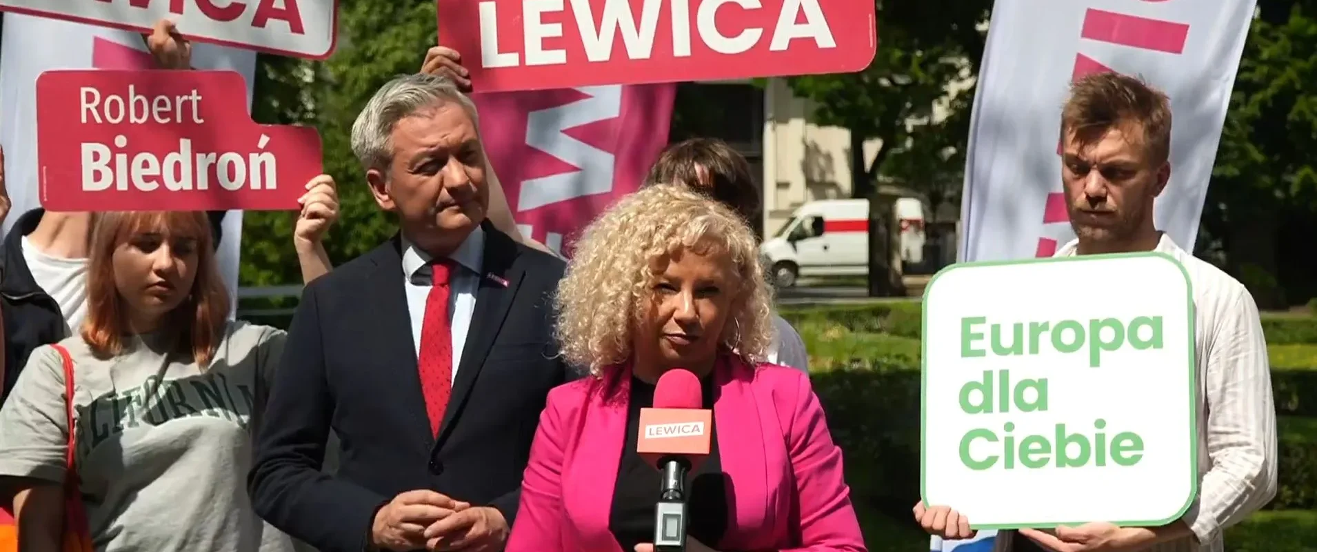 Kobieta o blond włosach w różowym żakiecie przemawia do mikrofonu podczas konferencji Lewicy, wokół niej widoczne są plakaty z napisem LEWICA i znak „Europa dla Ciebie”.