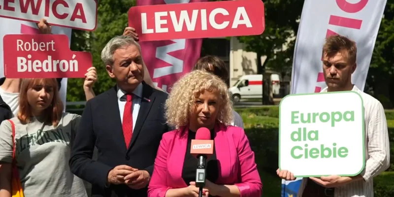 Kobieta o blond włosach w różowym żakiecie przemawia do mikrofonu podczas konferencji Lewicy, wokół niej widoczne są plakaty z napisem LEWICA i znak „Europa dla Ciebie”.