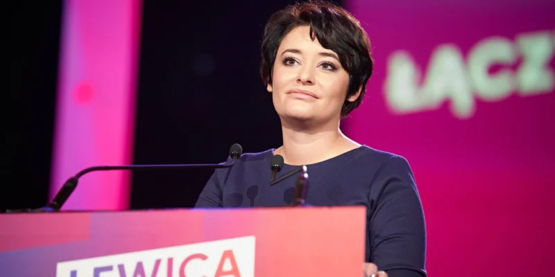 Kobieta z krótkimi czarnymi włosami przemawia z podiumem z napisem LEWICA podczas konferencji, stojąc przed mikrofonami na różowo-fioletowym tle.