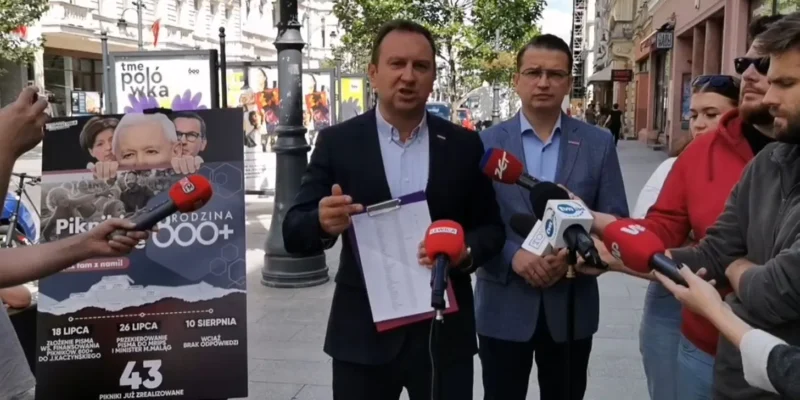 Mężczyzna w garniturze przemawia do mikrofonów podczas konferencji prasowej na ulicy; obok niego stoi drugi mężczyzna, a w tle widać plakat promujący Pikniki 800 Plus.