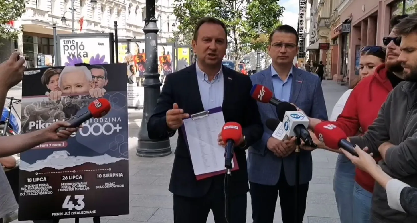 Mężczyzna w garniturze przemawia do mikrofonów podczas konferencji prasowej na ulicy; obok niego stoi drugi mężczyzna, a w tle widać plakat promujący Pikniki 800 Plus.