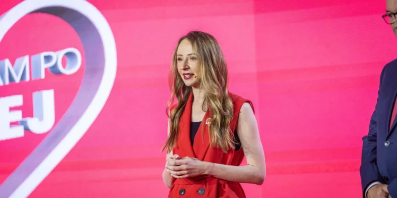 Kobieta na scenie, ubrana w czerwoną kamizelkę, przemawia do publiczności na tle różowego ekranu z informacją o 1500 zł na opłatę żłobkową.