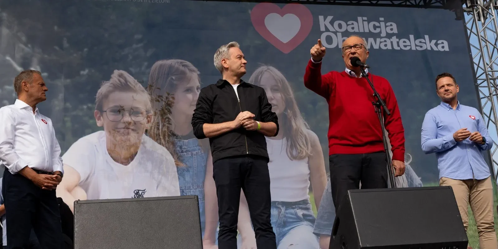 Grupa mężczyzn na scenie podczas Marszu Miliona Serc; przemawiający mężczyzna w czerwonym swetrze stoi przy mikrofonie, a w tle widnieje baner Koalicji Obywatelskiej i serce.