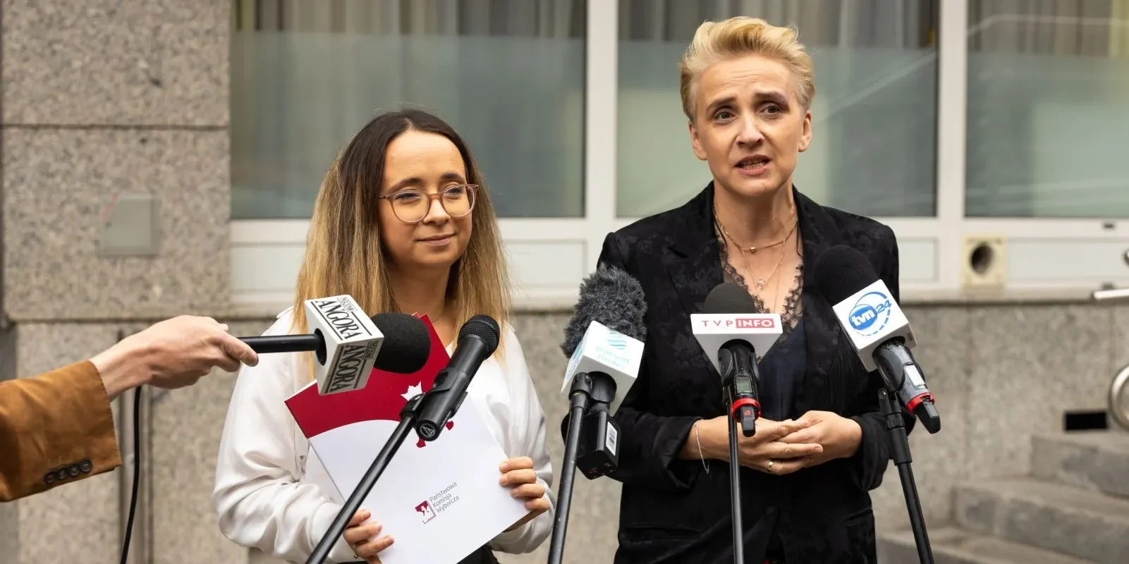 Dwie kobiety stoją przed mikrofonami na konferencji prasowej, jedna trzyma teczkę z dokumentami dotyczącymi rejestracji komitetu wyborczego Lewicy.