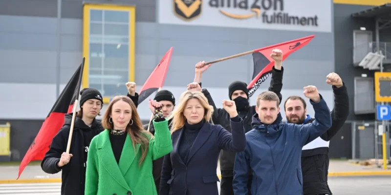 Grupa demonstrantów stoi przed budynkiem Amazon Fulfillment, unosząc pięści i trzymając flagi podczas protestu.