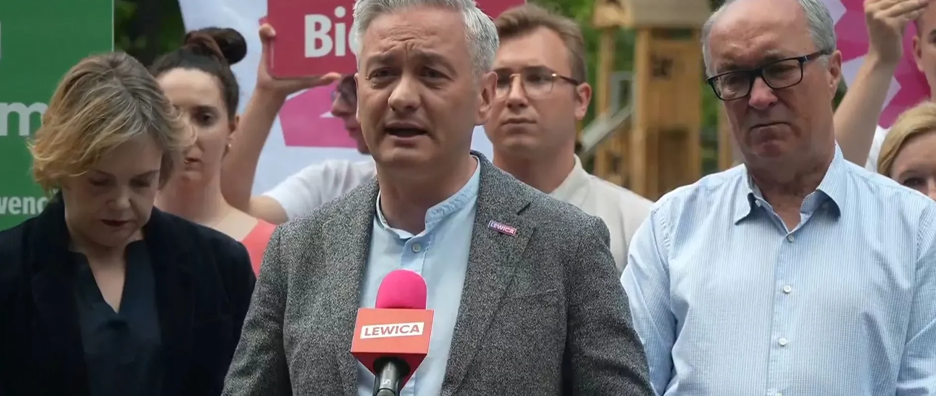 Grupa osób na wiecu w kontekście sytuacji na granicy; centralny mężczyzna w szarym garniturze przemawia do mikrofonu z logiem Lewicy, w otoczeniu innych uczestników i plakatów w tle.