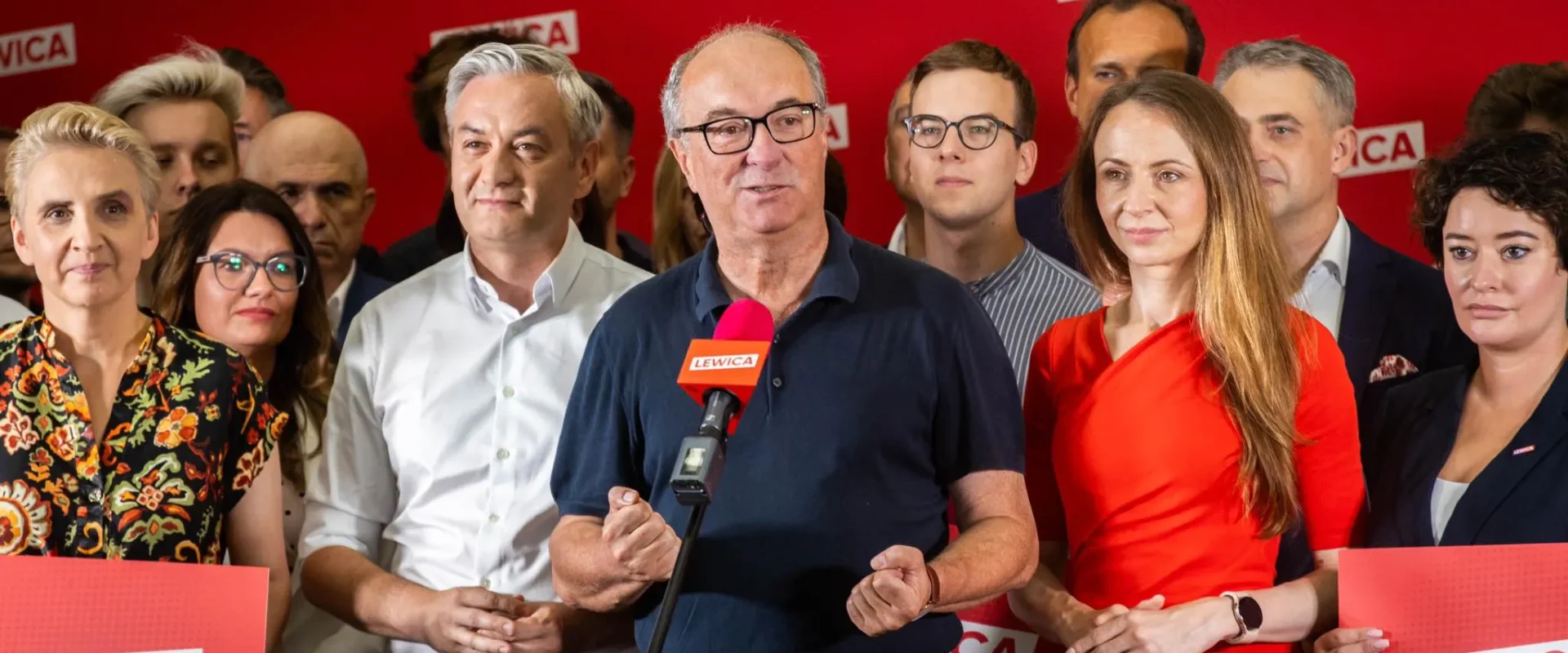 Grupa polityków stoi na scenie przed czerwonym tłem z logo Lewica; w centrum mężczyzna przemawia do mikrofonu z logiem LEWICA.
