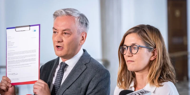 Mężczyzna w garniturze trzyma dokument w purpurowej teczce podczas konferencji prasowej, obok niego stoi kobieta z okularami.