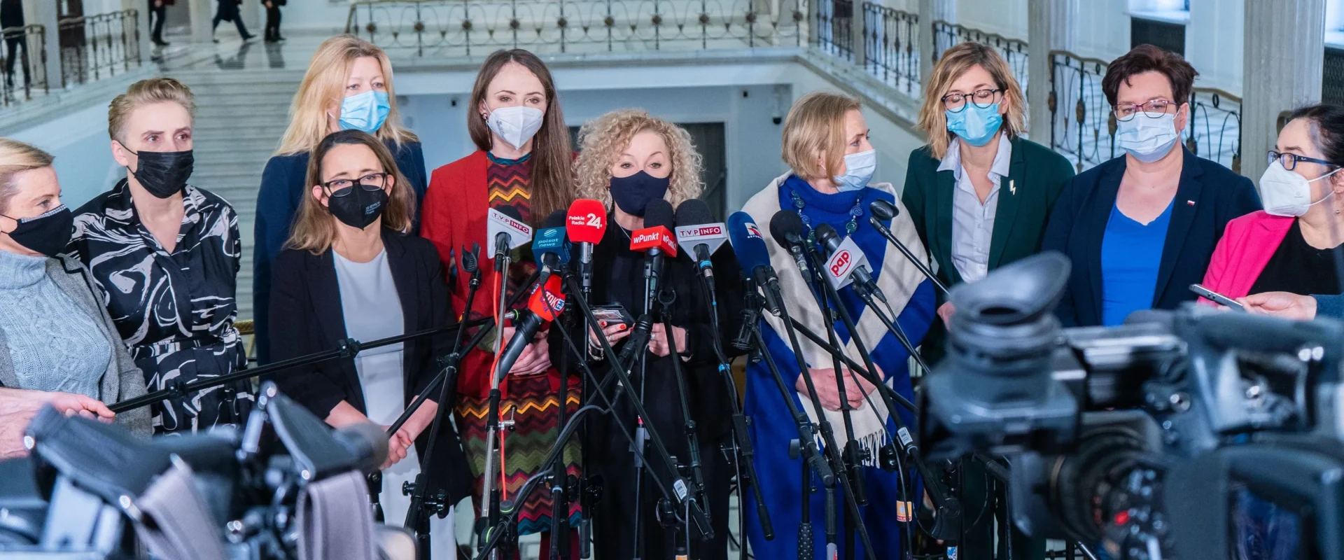 Grupa kobiet w masce ochronnej stoi przed mikrofonami w korytarzu budynku, prawdopodobnie podczas konferencji prasowej.