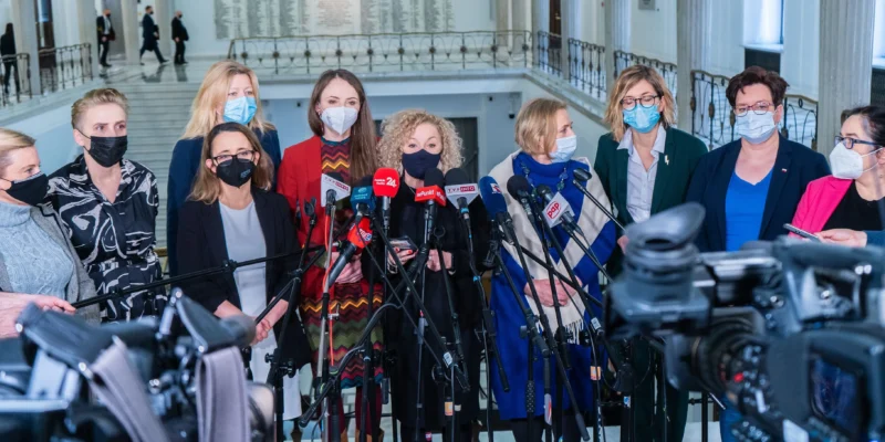 Grupa kobiet w masce ochronnej stoi przed mikrofonami w korytarzu budynku, prawdopodobnie podczas konferencji prasowej.