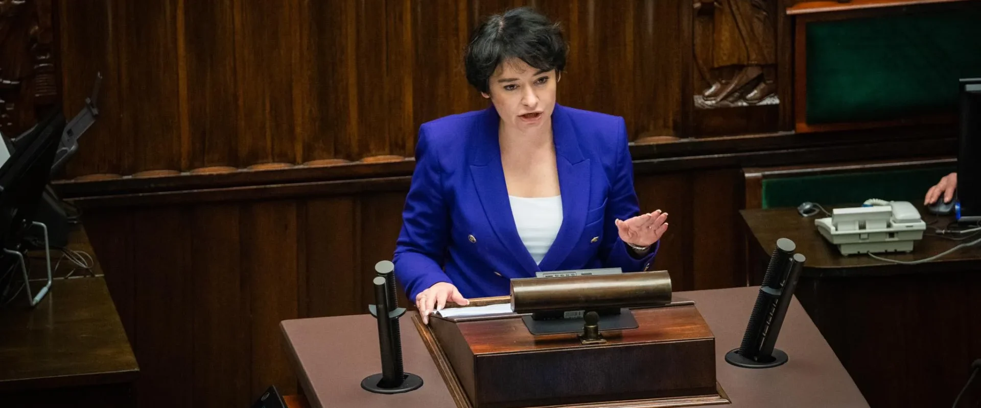 Kobieta w niebieskiej marynarce przemawia zza mównicy w sali plenarnej, otoczonej drewnianymi panelami.