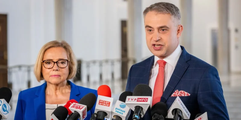 Dwóch polityków na konferencji prasowej w korytarzu, stojących przed licznymi mikrofonami; mężczyzna w granatowym garniturze z czerwonym krawatem przemawia, obok niego kobieta w niebieskim żakiecie.