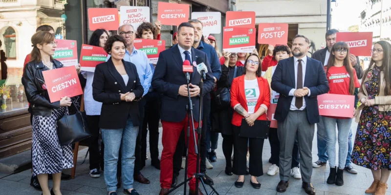 Grupa osób na ulicznym wiecu politycznym; w centrum mężczyzna w garniturze przemawia do mikrofonów, otoczony innymi uczestnikami trzymającymi czerwone plakaty Lewica.