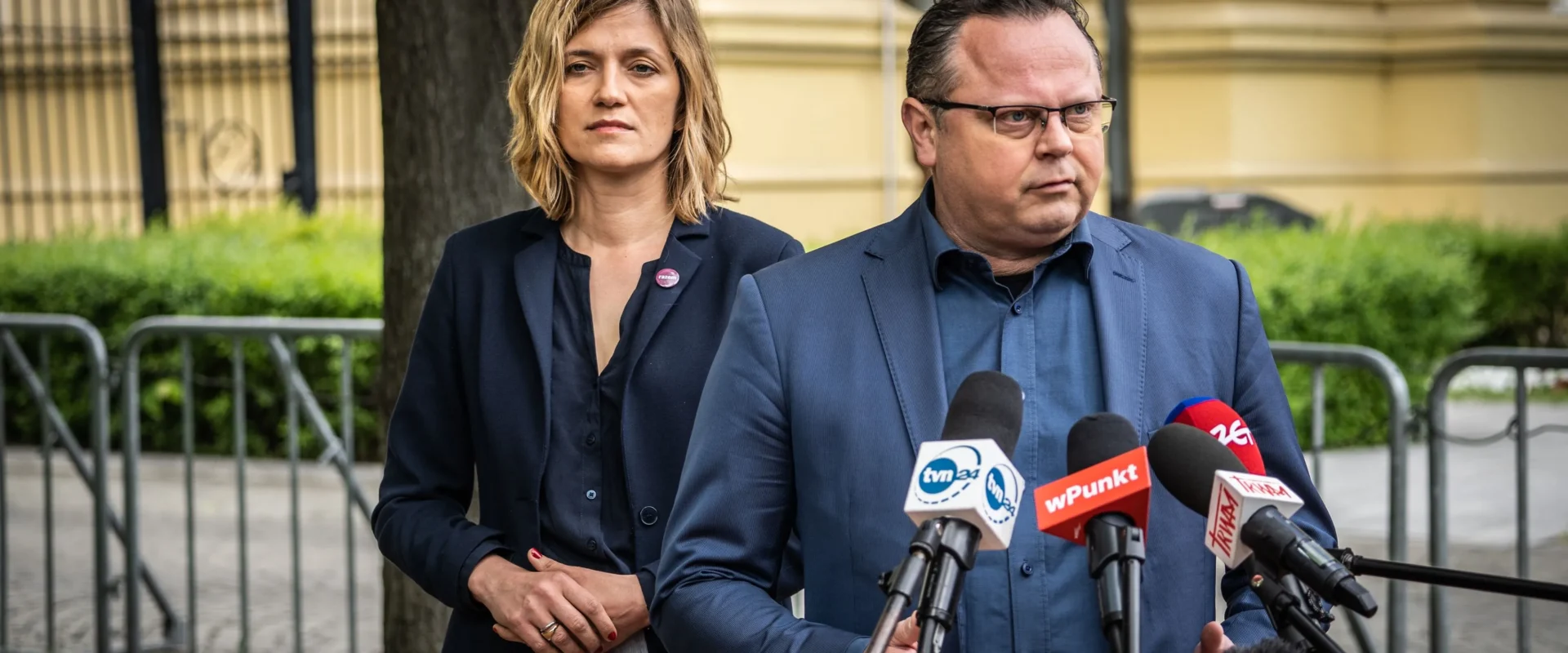 Dwóch polityków stoi na zewnątrz przed zestawem mikrofonów podczas konferencji prasowej; mężczyzna w granatowym garniturze przemawia, a kobieta stoi obok.