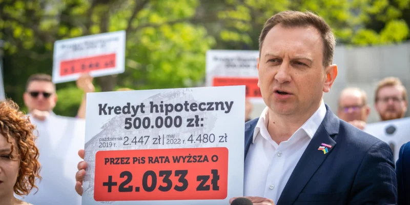 Na zdjęciu widoczny jest mężczyzna mówiący do mikrofonu podczas protestu, trzymający transparent informujący o wzroście raty kredytu hipotecznego o 2.033 zł.
