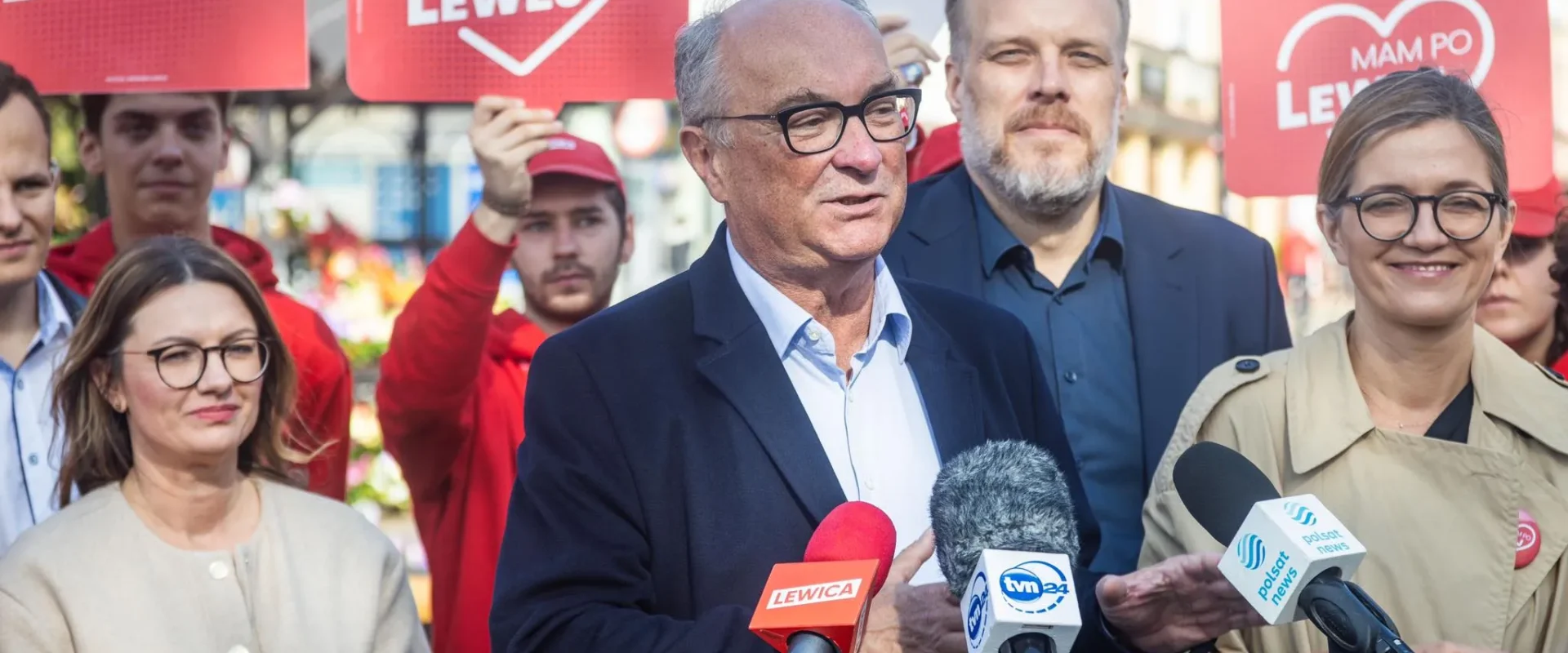 Osoba przemawia do mikrofonów podczas konferencji prasowej Lewicy, otoczona przez zwolenników i czerwone plakaty z napisem LEWICA.