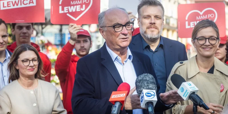 Osoba przemawia do mikrofonów podczas konferencji prasowej Lewicy, otoczona przez zwolenników i czerwone plakaty z napisem LEWICA.