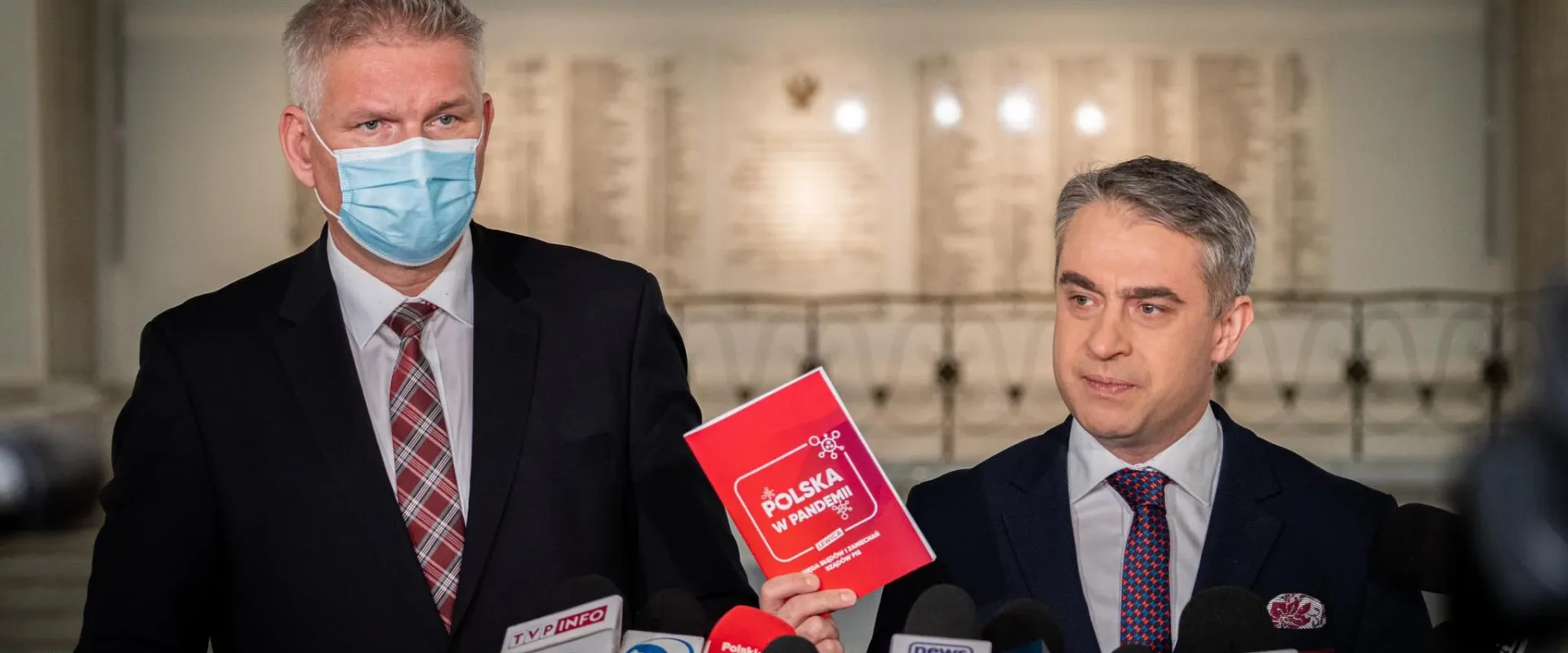 Na zdjęciu dwóch mężczyzn w garniturach, jeden z nich w niebieskiej masce, prezentuje publikację zatytułowaną "Polska w pandemii" podczas konferencji prasowej.