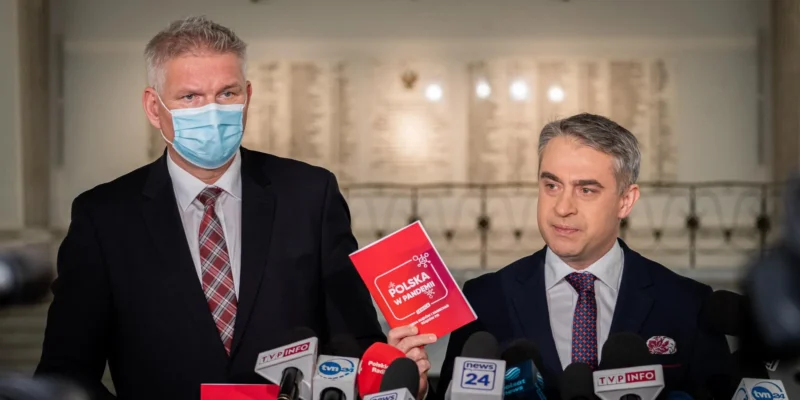 Na zdjęciu dwóch mężczyzn w garniturach, jeden z nich w niebieskiej masce, prezentuje publikację zatytułowaną "Polska w pandemii" podczas konferencji prasowej.