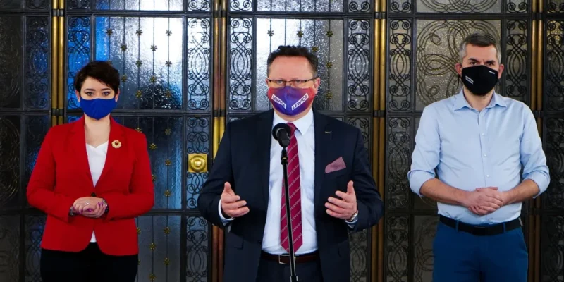 Trzy osoby stoją przed ozdobną przeszkloną ścianką; kobieta w czerwonej marynarce, mężczyzna w garniturze przy mikrofonie, oraz drugi mężczyzna w jasnej koszuli, wszyscy w maseczkach.