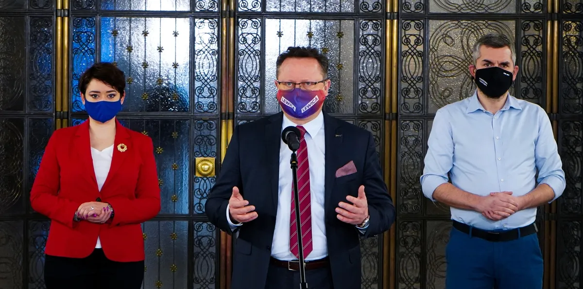 Trzy osoby stoją przed ozdobną przeszkloną ścianką; kobieta w czerwonej marynarce, mężczyzna w garniturze przy mikrofonie, oraz drugi mężczyzna w jasnej koszuli, wszyscy w maseczkach.