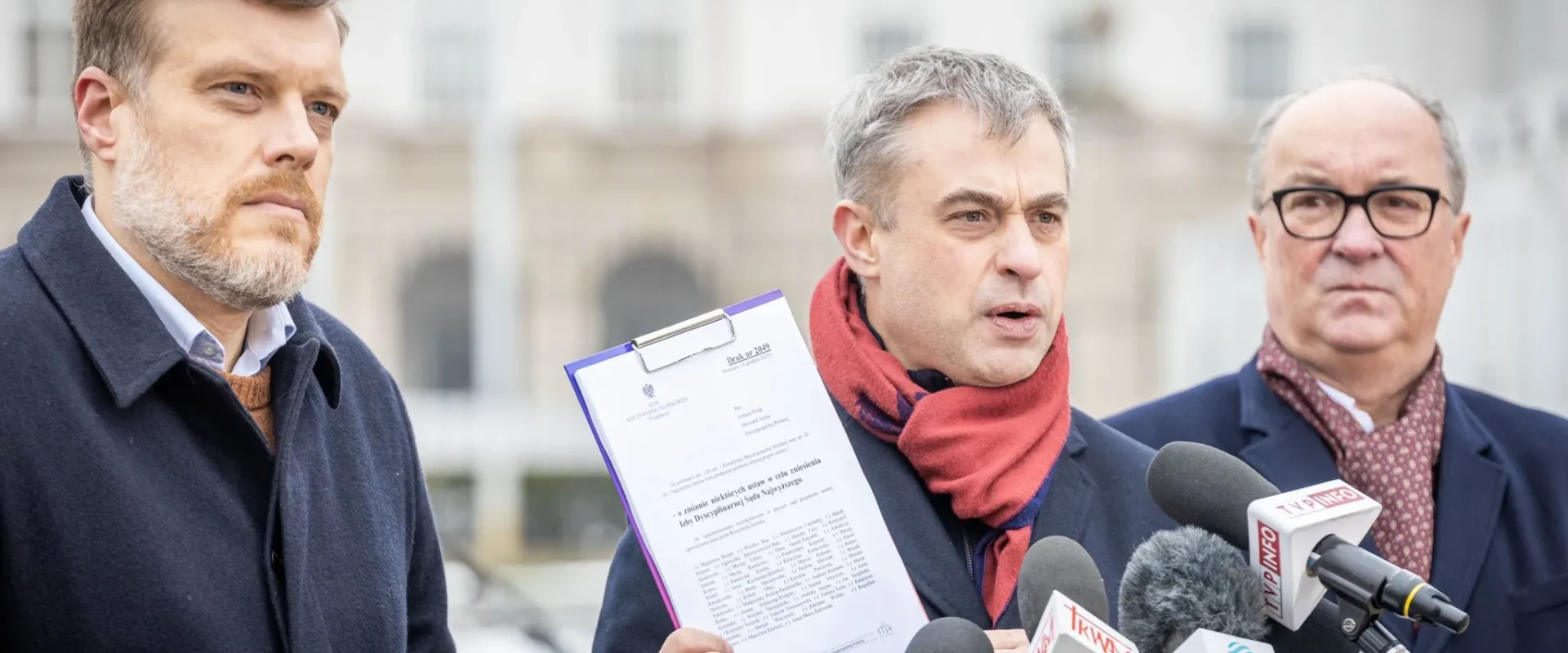 Trzech mężczyzn w garniturach podczas konferencji prasowej na zewnątrz, z których jeden trzyma kartę z dokumentem, a przed nimi stoją liczne mikrofony z logotypami mediów.