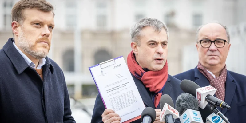 Trzech mężczyzn w garniturach podczas konferencji prasowej na zewnątrz, z których jeden trzyma kartę z dokumentem, a przed nimi stoją liczne mikrofony z logotypami mediów.