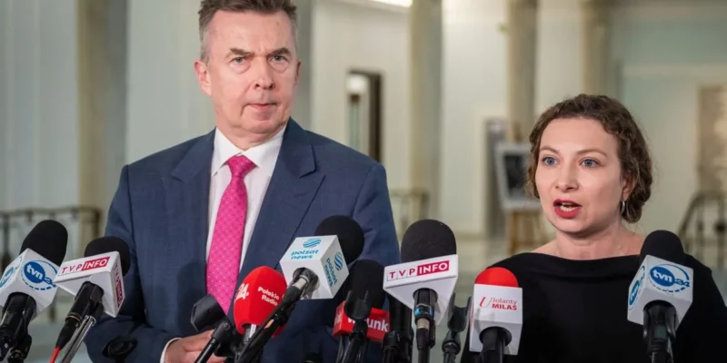 Na zdjęciu widoczna jest dwójka polityków podczas konferencji prasowej, w otoczeniu mikrofonów ze znakami różnych mediów.