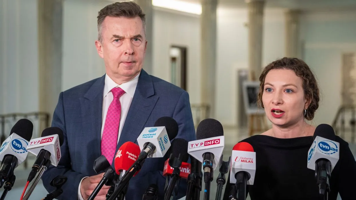 Na zdjęciu widoczna jest dwójka polityków podczas konferencji prasowej, w otoczeniu mikrofonów ze znakami różnych mediów.