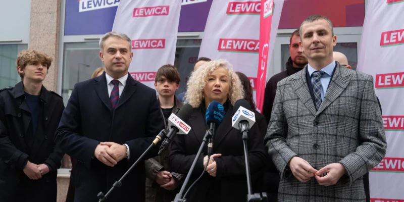 Grupa osób podczas konferencji prasowej Lewicy, stojących przed mikrofonami na tle flag z napisem LEWICA.