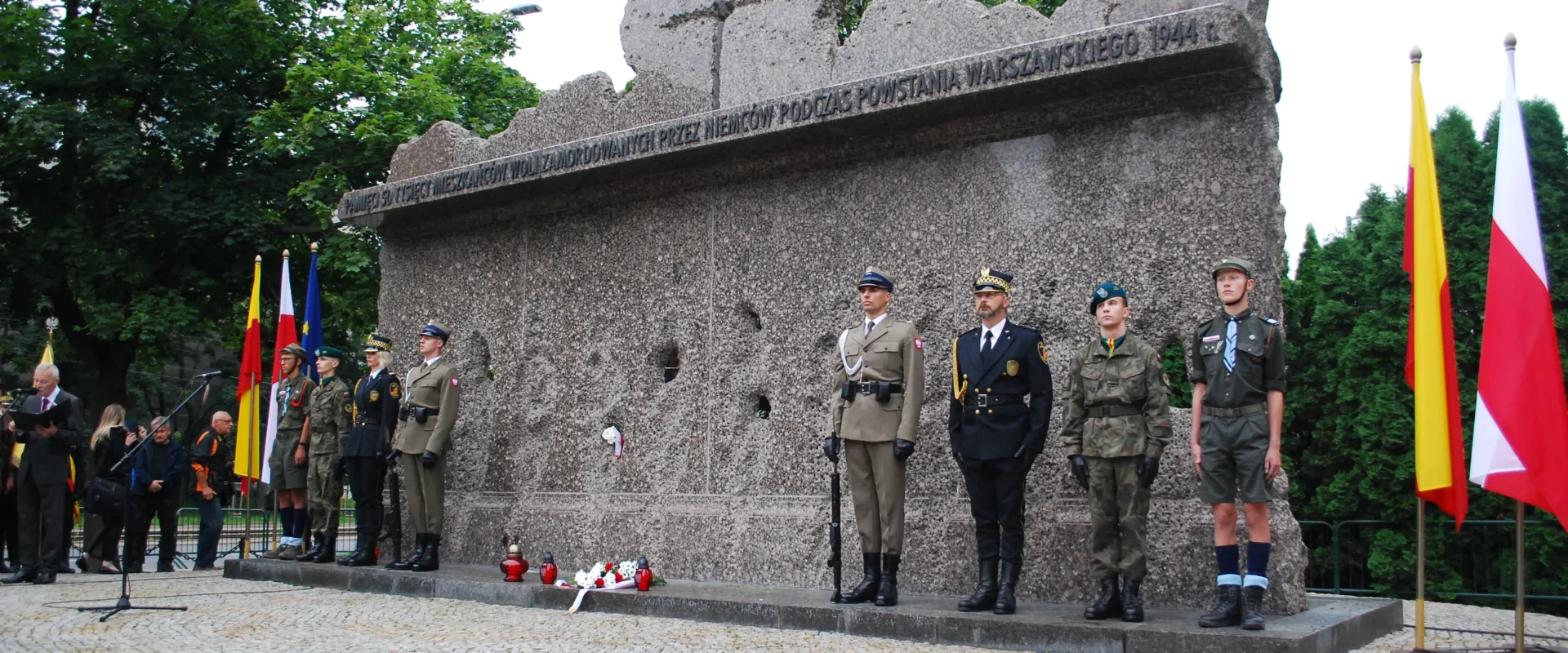Uczestnicy w mundurach składają hołd ofiarom Rzezi Woli podczas uroczystości upamiętniającej 77. rocznicę tragedii, stojąc przed monumentalnym pomnikiem z inskrypcją i flagami po bokach.
