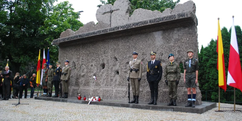 Uczestnicy w mundurach składają hołd ofiarom Rzezi Woli podczas uroczystości upamiętniającej 77. rocznicę tragedii, stojąc przed monumentalnym pomnikiem z inskrypcją i flagami po bokach.