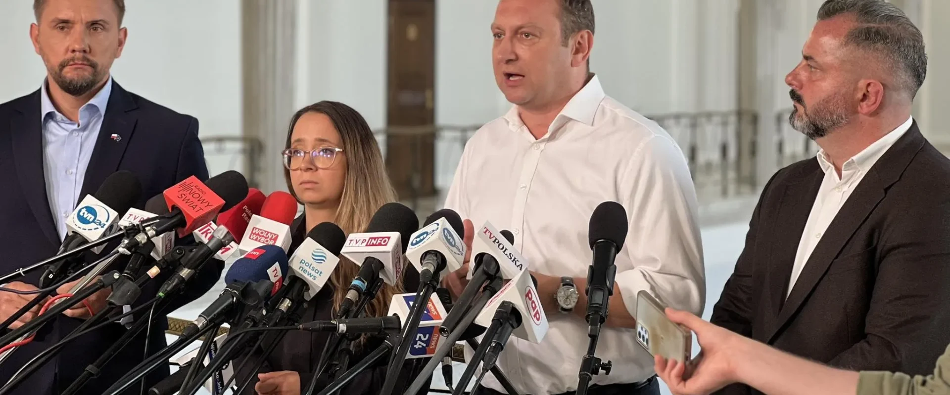 Grupa osób podczas konferencji prasowej stoi przed licznymi mikrofonami mediów, a mężczyzna w białej koszuli wygłasza oświadczenie.