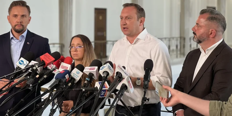 Grupa osób podczas konferencji prasowej stoi przed licznymi mikrofonami mediów, a mężczyzna w białej koszuli wygłasza oświadczenie.