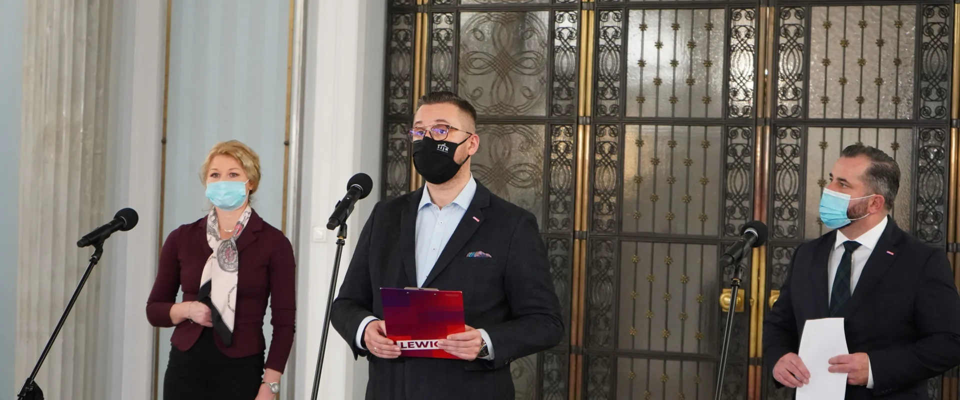 Trzech polityków w maseczkach stoi przy mikrofonach podczas konferencji prasowej w Sejmie, a w tle widoczne są ozdobne drzwi; środkowy mężczyzna trzyma folder z napisem Lewica.